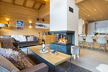 Cozy wooden chalet interior featuring a modern three-sided fireplace, plush sofas, and a dining area in the background.