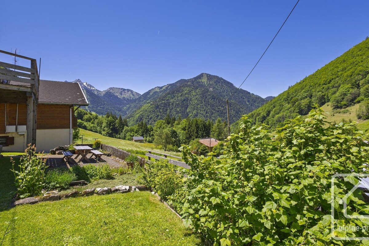 A cozy house with a picnic area in a lush green valley, surrounded by majestic, forest-covered mountains under a clear blue sky.
