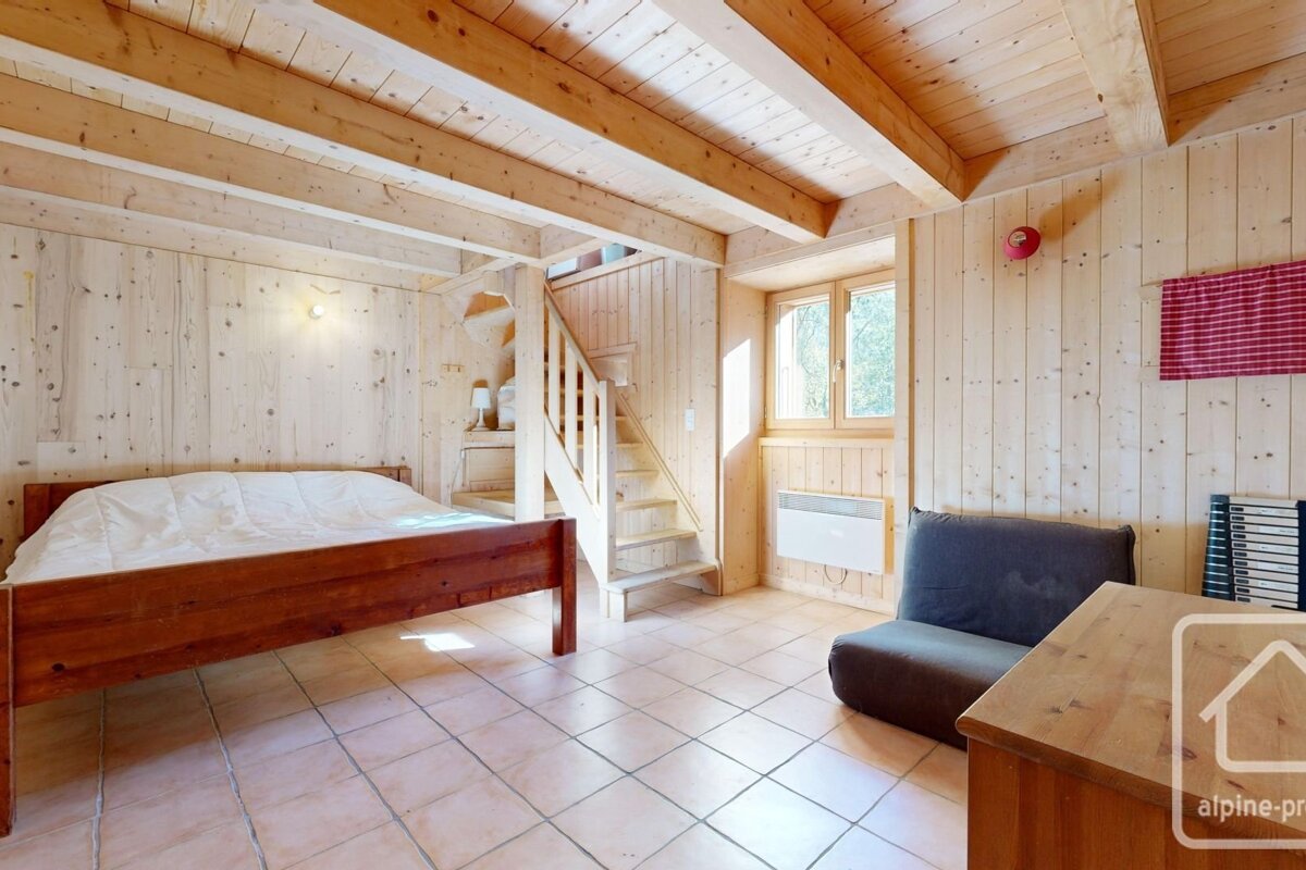 Rustic room with light wood walls and ceiling beams. Features a wooden bed, tiled floor, an open staircase, a window, and a dark sofa.
