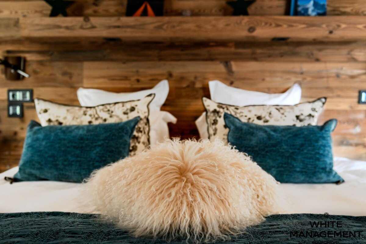 A cozy bed styled with a fluffy beige pillow, teal cushions, and patterned pillows against a rustic wooden wall.