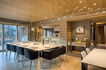 A luxurious dining room featuring a long set table, unique suspended lighting, and wooden walls. Large windows offer a view of snowy trees outside.