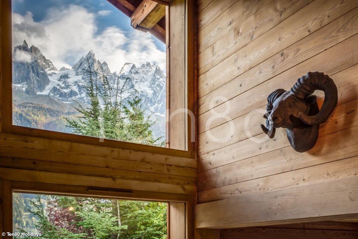 A cozy wooden interior frames a stunning view of snow-capped mountains and green trees. A decorative ram's head adorns the wood-paneled wall.