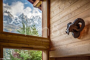 A cozy wooden interior frames a stunning view of snow-capped mountains and green trees. A decorative ram's head adorns the wood-paneled wall.
