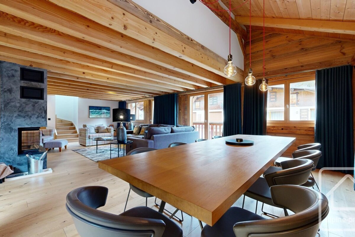 A spacious, modern open-plan living/dining area featuring wooden beams, light wood floors, a contemporary fireplace, and a long dining table.
