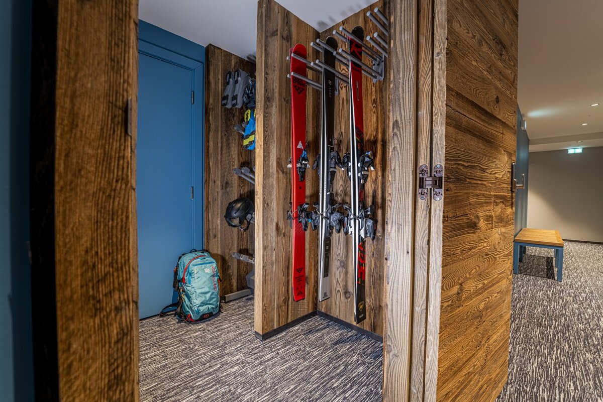 A rustic ski storage area with wood paneling, skis, boots, helmet, and a backpack. A blue door and textured carpet are visible.