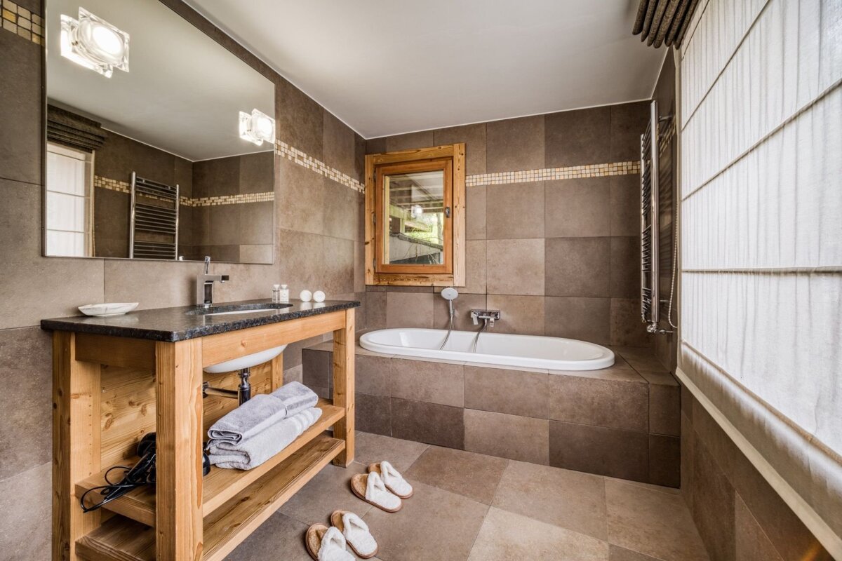 A rustic brown-tiled bathroom with a wooden vanity, granite countertop, built-in tub, large mirror, and window. Slippers are on the tiled floor.
