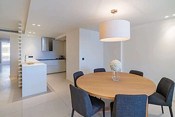 A white, modern open-concept kitchen flows into a dining area with a round wood table, dark chairs, and a large drum pendant light.