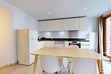 A modern, minimalist kitchen and dining area featuring white cabinetry, black countertops, a white fridge, and a light wood table with white chairs.