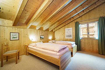 A cozy wooden bedroom with a slanted beamed ceiling, two twin beds with pink duvets, and a window with green curtains.