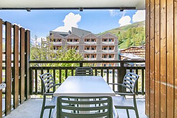 Wooden balcony with a table and chairs overlooking scenic resort buildings and green hills under a blue sky.