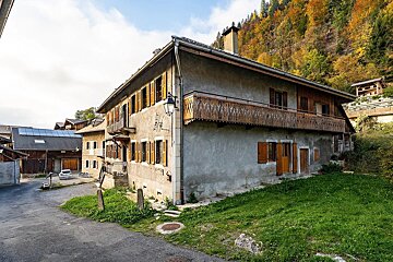 An old, gray house with wooden shutters and a carved balcony in a mountain village, surrounded by colorful autumn trees and green grass.