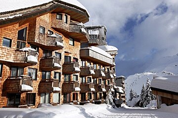 Modern white ski resort buildings with balconies cascade down a mountain slope, surrounded by bare trees, snow patches, and traditional chalets.