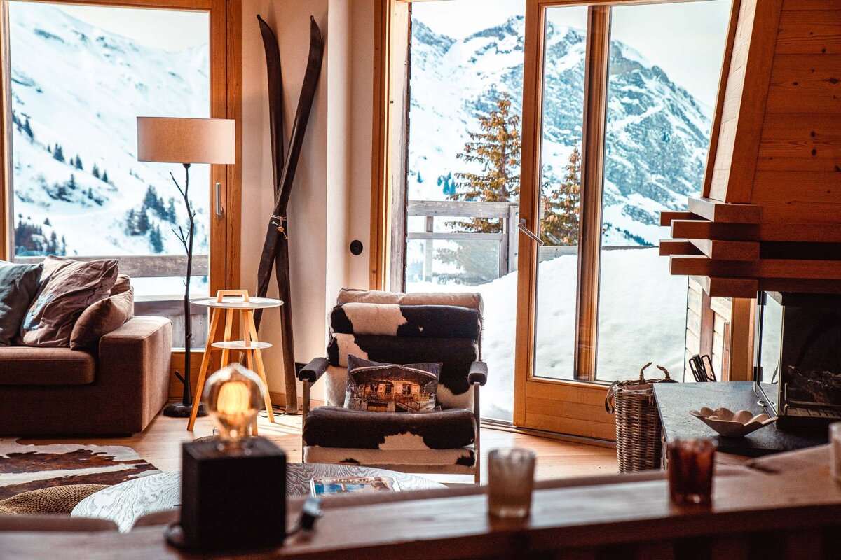Cozy ski chalet interior with a cowhide chair and large windows offering a breathtaking view of snowy mountains.
