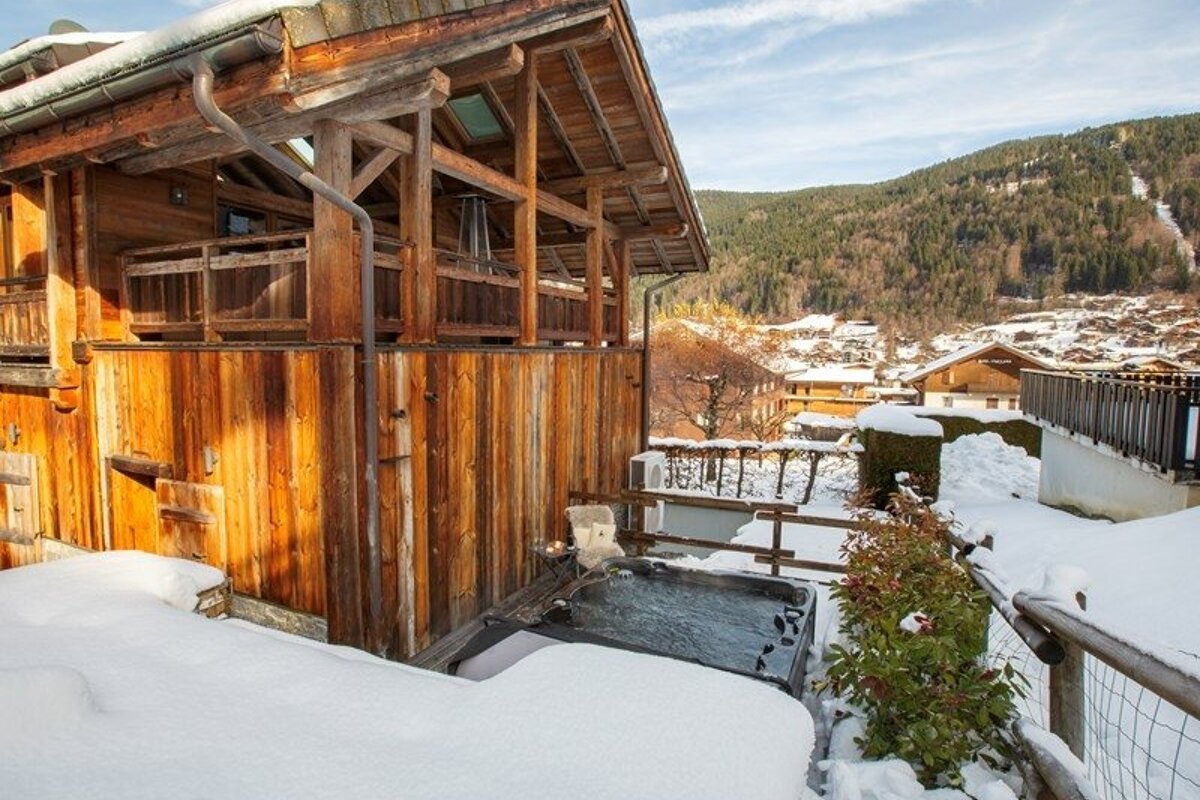 A cozy wooden chalet with an outdoor hot tub surrounded by snow, overlooking a mountain village in winter.