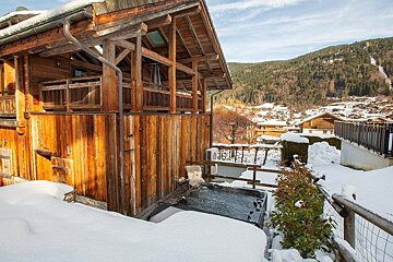A cozy wooden chalet with an outdoor hot tub surrounded by snow, overlooking a mountain village in winter.