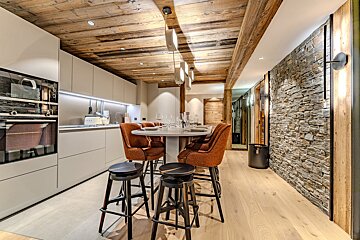 Chic chalet-style kitchen & dining area with a rustic wooden ceiling, stone accent wall, modern light gray cabinets, and inviting orange chairs.