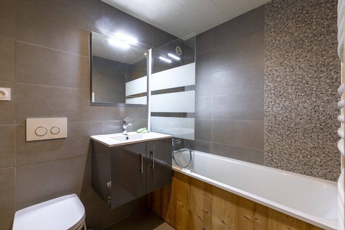 Modern grey tiled bathroom with a wood-paneled bathtub, vanity, mirror, and toilet.