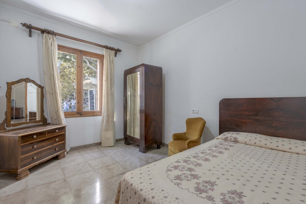 A classic-style bedroom with wooden furniture (dresser, wardrobe, bed), a patterned bedspread, tiled floor, and a window looking out to trees.
