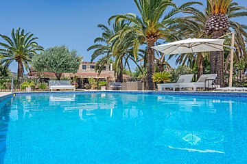 A vibrant blue swimming pool in the foreground, with lush palm trees, sun loungers, and a traditional stone villa under a clear sky.
