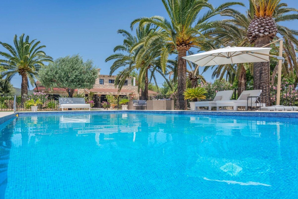 A sunny scene featuring a sparkling blue swimming pool, lush palm trees, white loungers, an umbrella, and a charming stone villa.