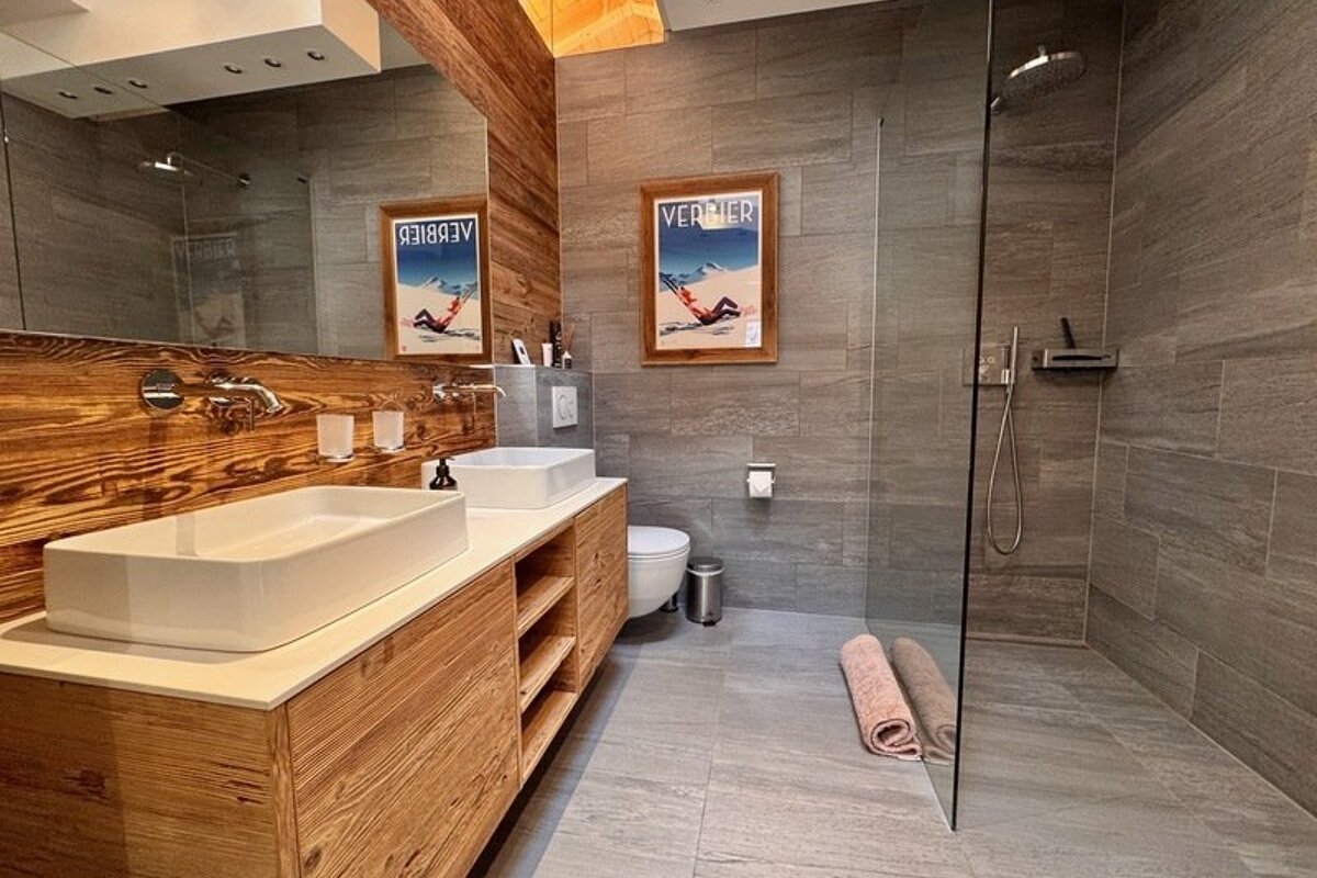 Modern rustic bathroom with gray tiling, a warm wooden vanity with double sinks, a toilet, and a walk-in glass shower.