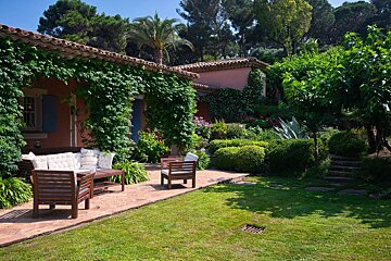 Sunny view of a vine-covered pink house with a tiled roof, blue shutters, and wooden patio furniture in a lush, manicured garden.