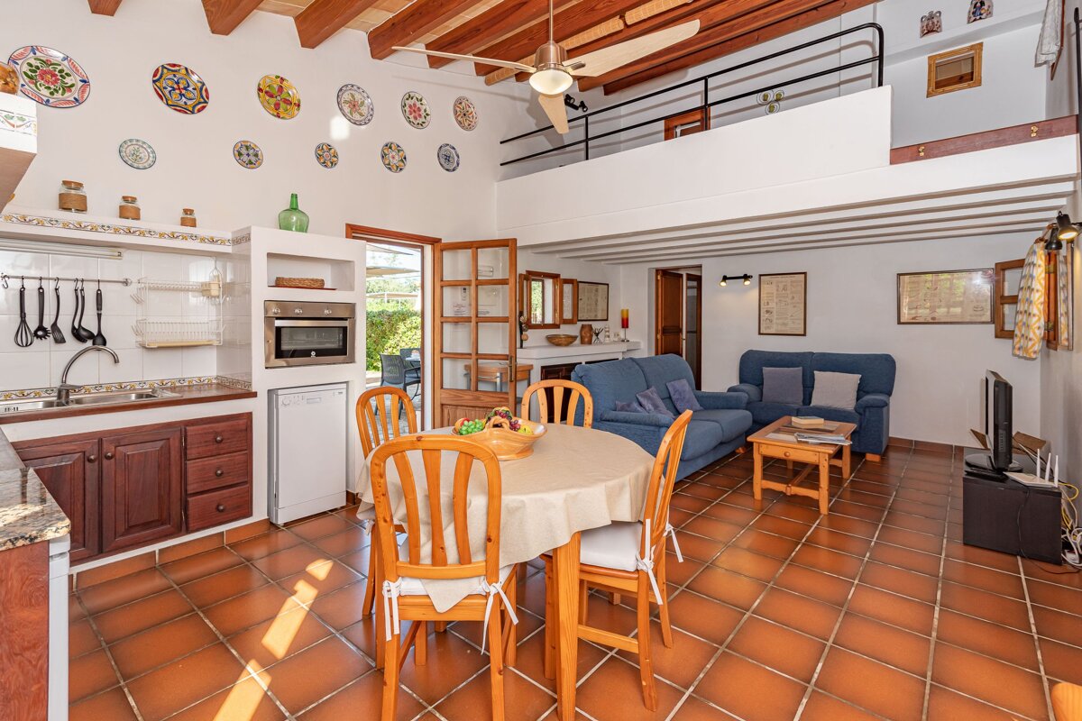 An open-plan room with a kitchen, dining area, and living room featuring blue sofas. It has terracotta floor tiles, wooden ceiling beams, and a mezzanine.