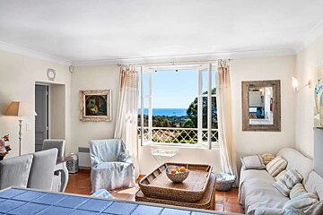 A bright living room with an open window revealing a beautiful blue ocean view and green landscape. Cozy furniture and natural light fill the space.