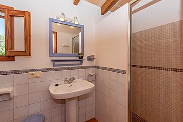 A bright bathroom features a white pedestal sink, blue framed mirror with lights, and a shower with brown mosaic tiles. It has white walls with a decorative tile border and a wooden-framed window.