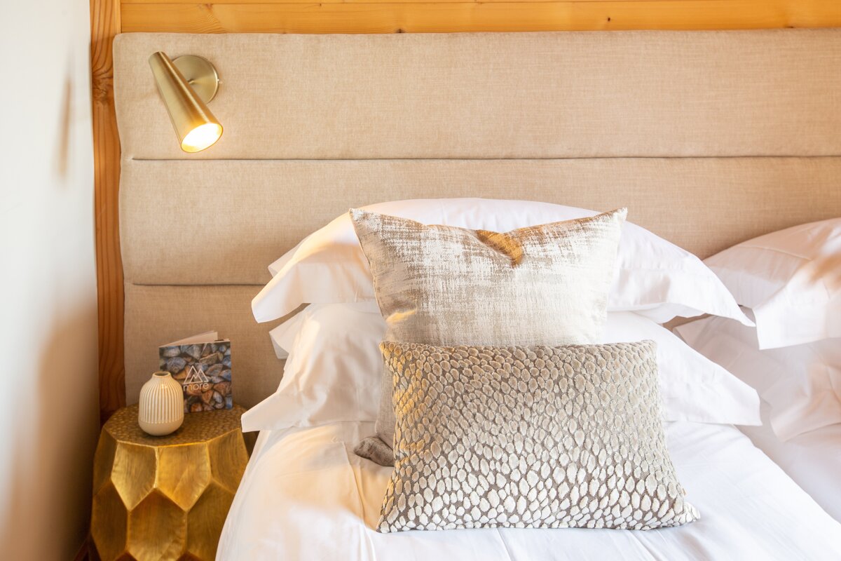 A luxurious bed with white linens, decorative pillows, a beige padded headboard, a gold sconce light, and a golden side table.