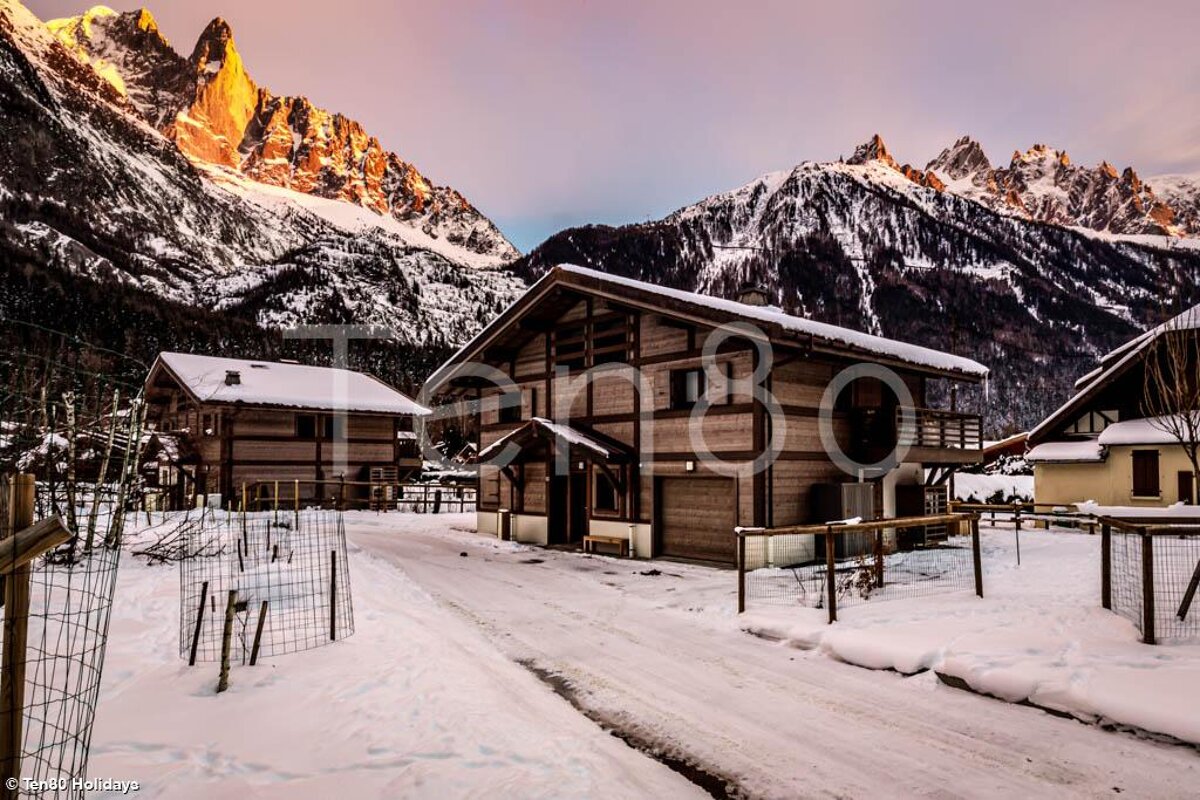 Snow-covered wooden chalets nestle before majestic, sun-kissed mountains, creating a picturesque winter scene at dawn or dusk.