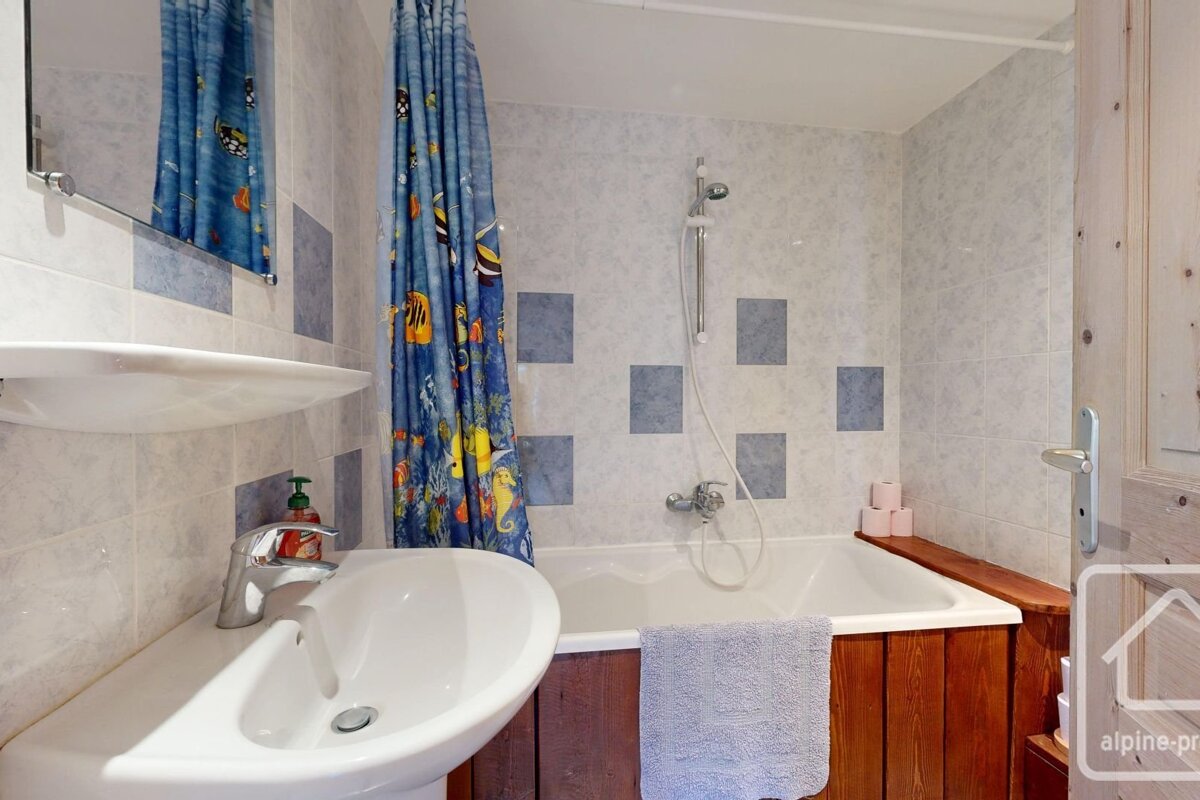 A bathroom with a white sink, a bathtub with wood paneling, a blue shower curtain with a fish pattern, and tiled walls.