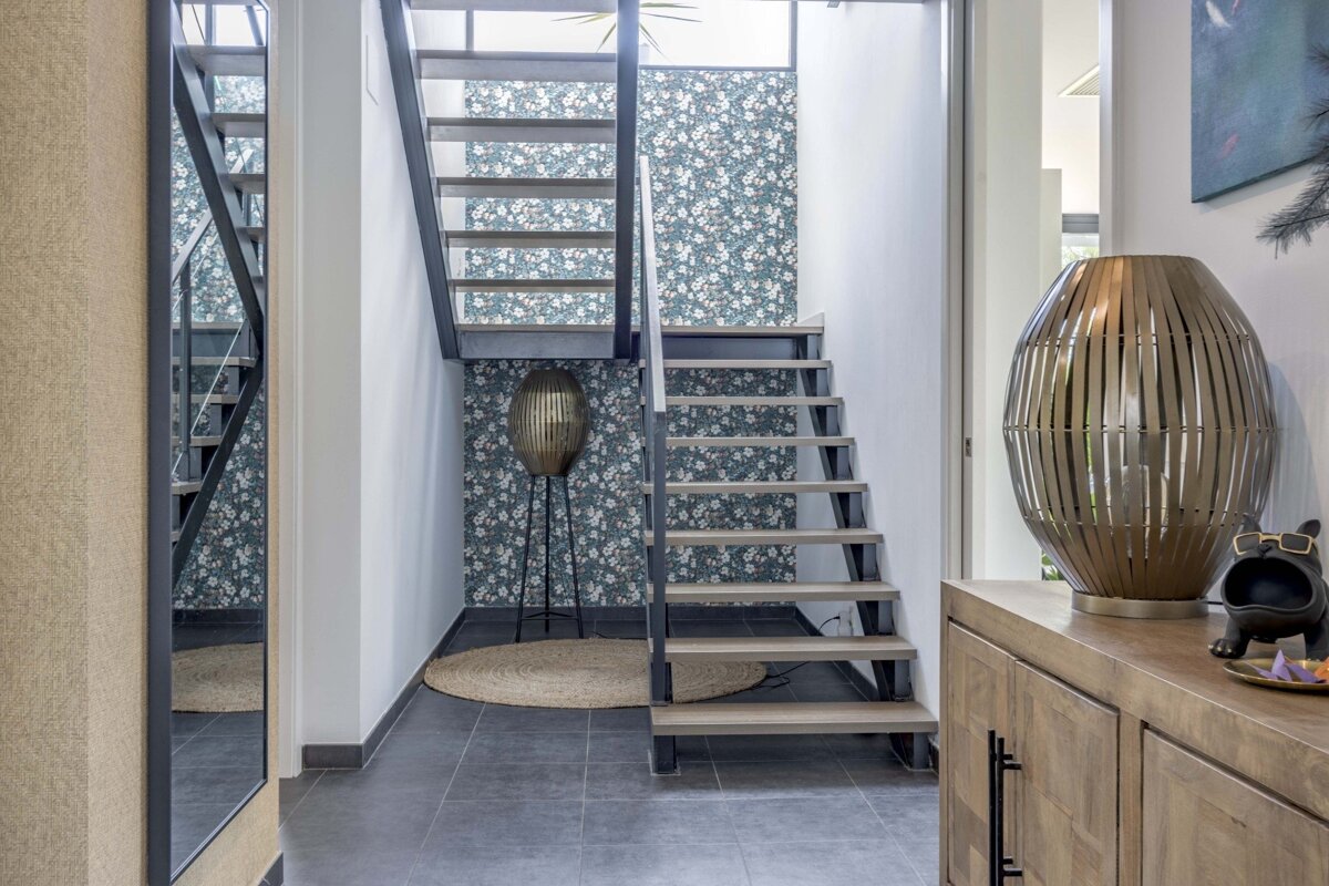 Modern entrance hall with an open staircase against blue floral wallpaper, featuring a metallic floor lamp and a wooden cabinet.