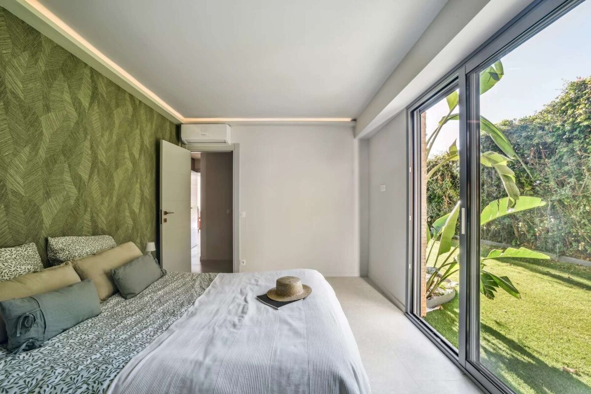 Bright bedroom with a green accent wall, large bed, and floor-to-ceiling glass doors looking out to a sunny, green garden.