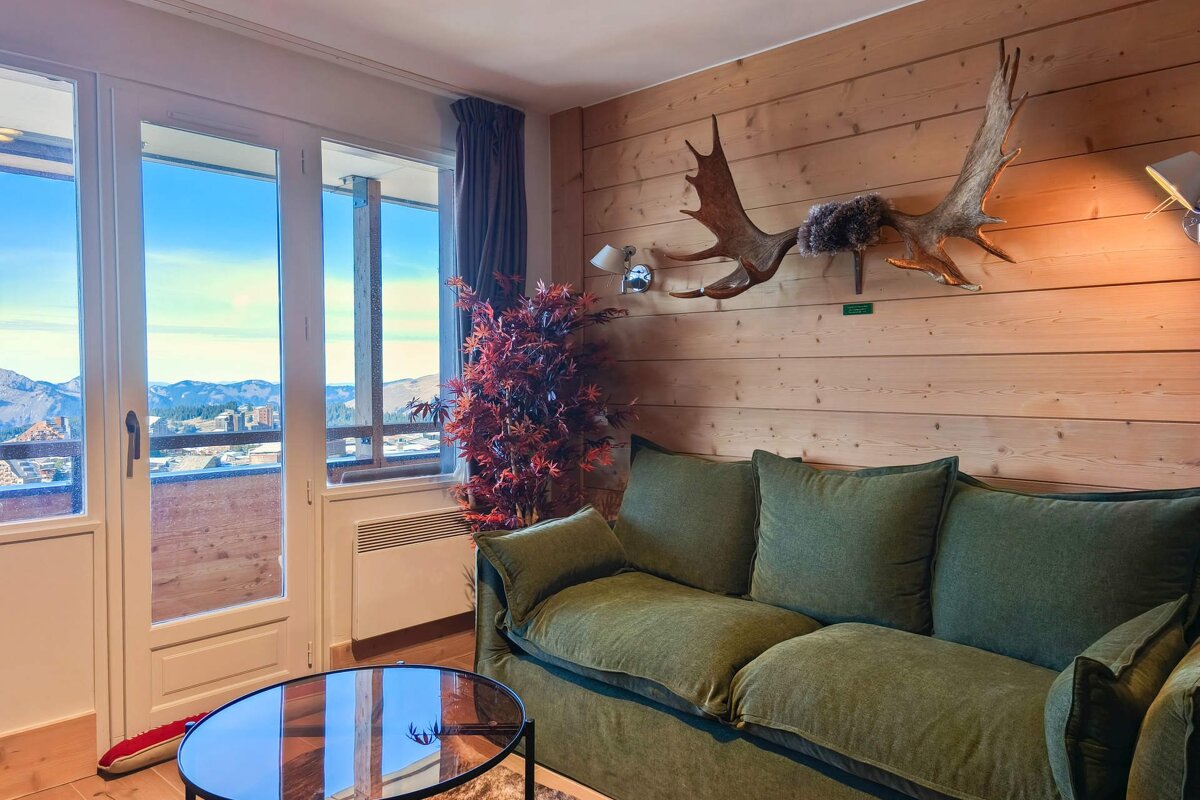Cozy chalet living room with mountain views, green sofa, wood-paneled wall, and large antlers decor.