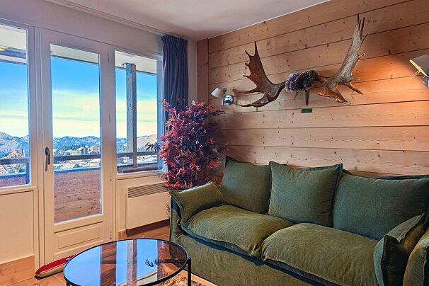 A cozy, wood-paneled living room features a beige sofa with two large teddy bears, a red shag rug, and a wooden coffee table. Heart decorations adorn the wall.