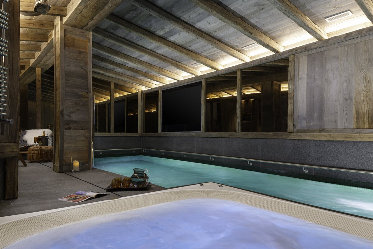 A rustic chalet indoor spa featuring a long swimming pool and a bubbling hot tub. Wooden beams, dark walls, and soft lighting create a serene atmosphere.