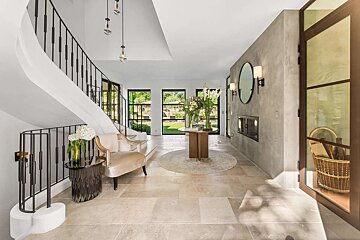 Bright, modern foyer featuring a curved staircase, stone floors, large windows with garden views, and a stylish central table.