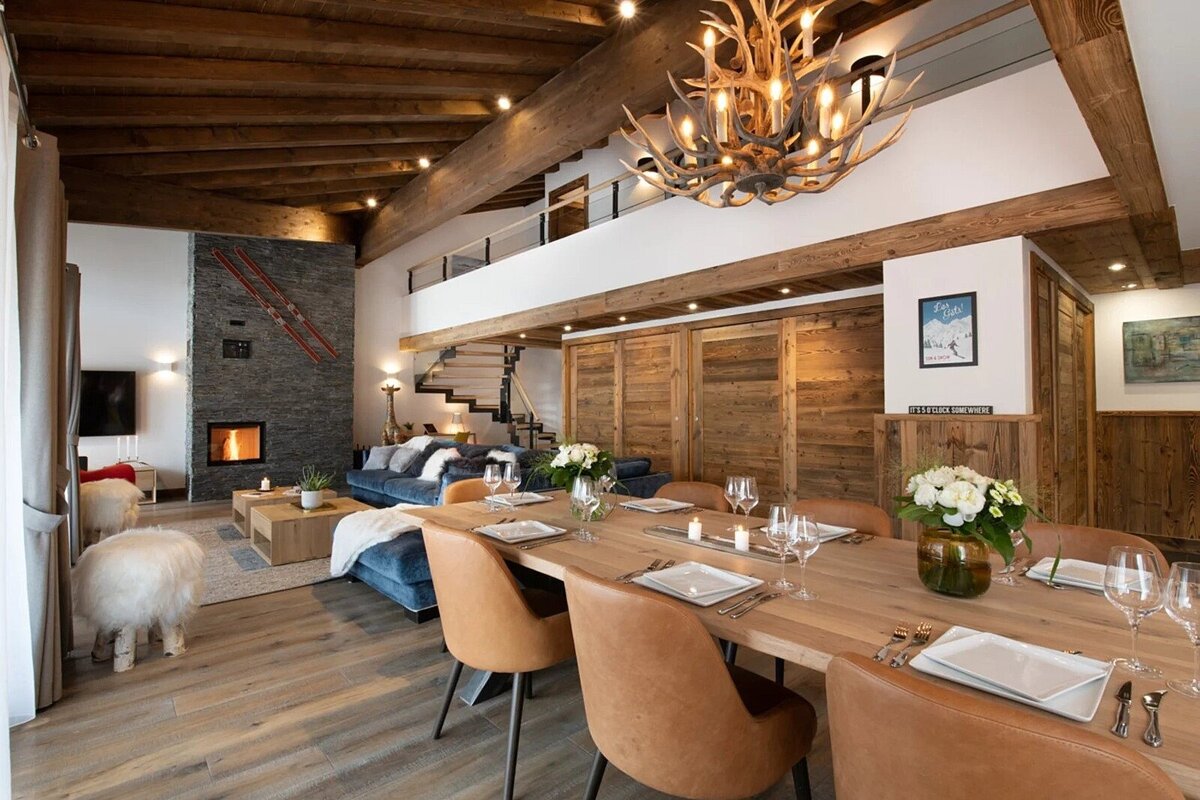 A luxurious, rustic chalet interior with an open-plan living and dining area, antler chandelier, stone fireplace, and wooden accents.