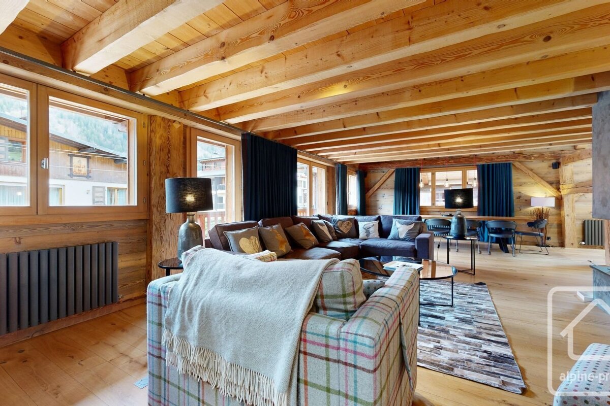 Cozy chalet living room with wooden walls and beamed ceiling. Features an L-shaped sofa, plaid armchair, and large windows with outdoor views.