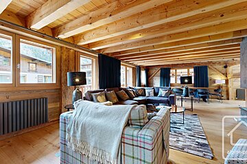 A cozy, rustic living room with extensive wood paneling and a beamed ceiling. Features a large sectional sofa, plaid armchair, and bright windows.