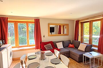 A sunny living/dining room features a grey sectional sofa, set white dining table, and large windows with red curtains overlooking trees. Wooden accents.