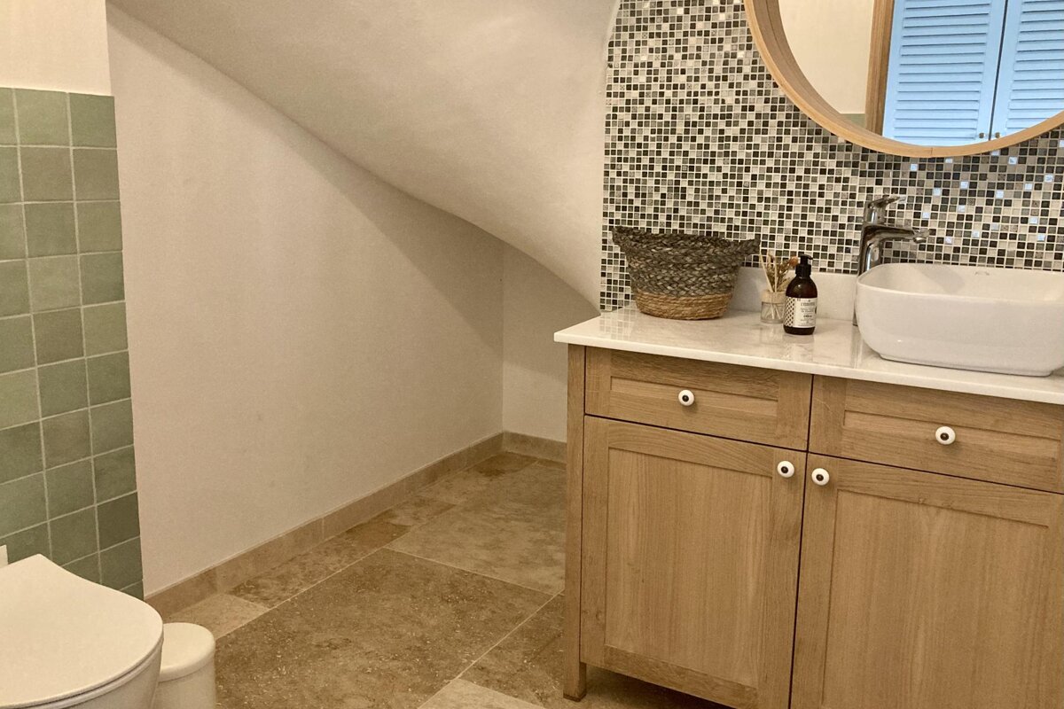 An attic bathroom with a light wood vanity, white basin, mosaic tile backsplash, round mirror, and sage green wall tiles.