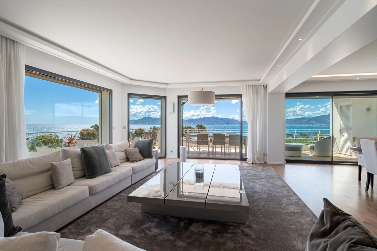 A bright, modern living room featuring a long sofa and large coffee table, with expansive windows offering panoramic ocean and mountain views.