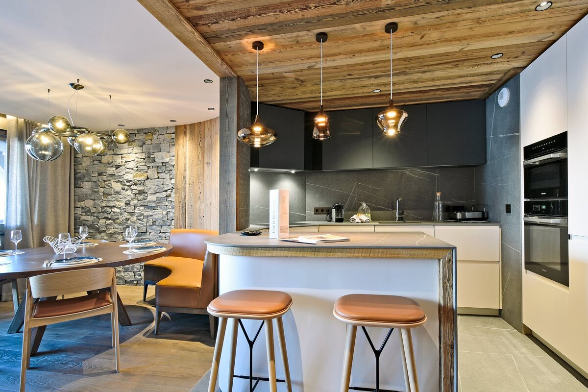 Modern chalet kitchen and dining area with warm wood ceilings, a stone accent wall, sleek cabinetry, and elegant pendant lights.
