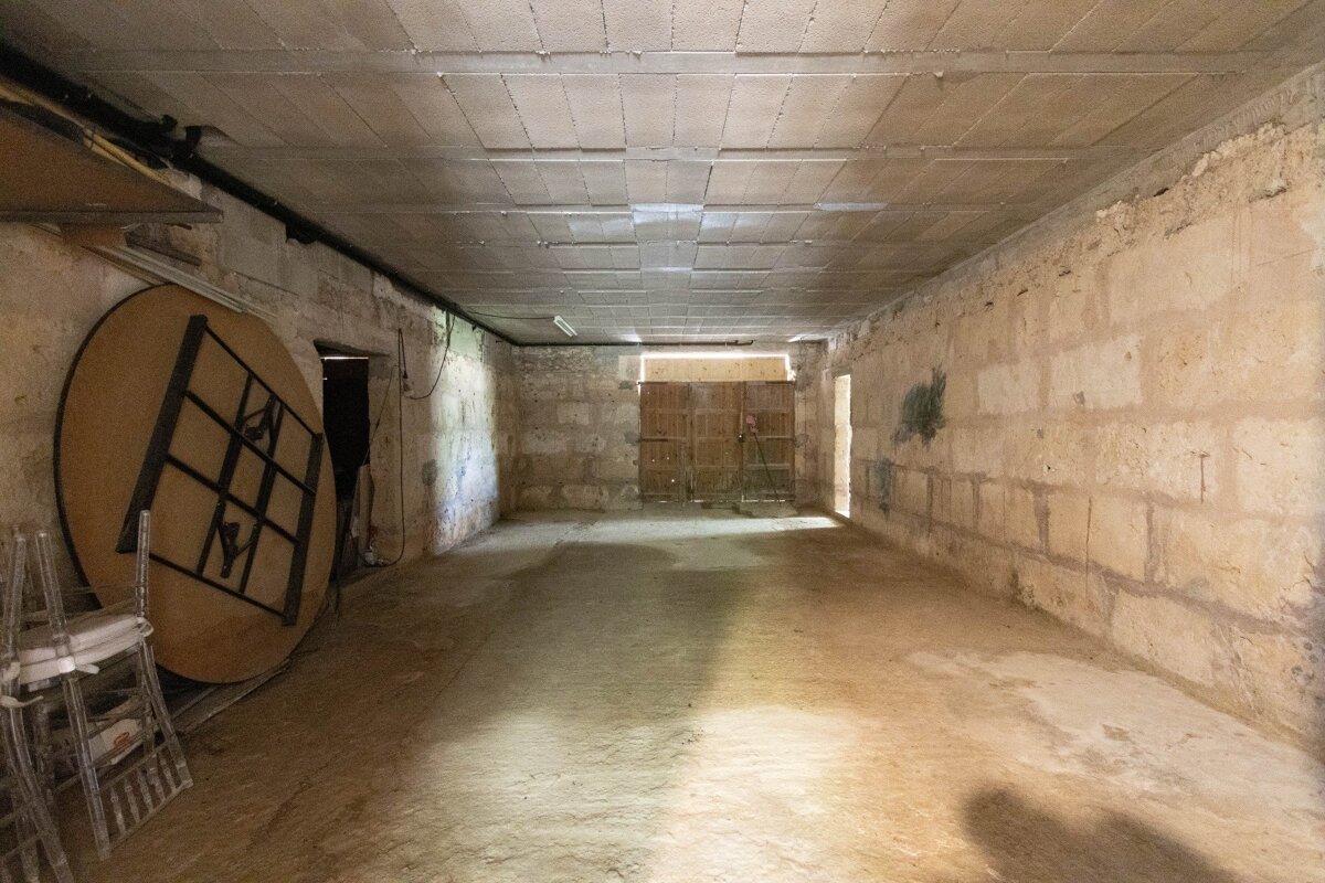 An empty, long room with rustic stone walls, concrete floor, and a tiled ceiling. A large round table leans left, and double wooden doors are at the far end.