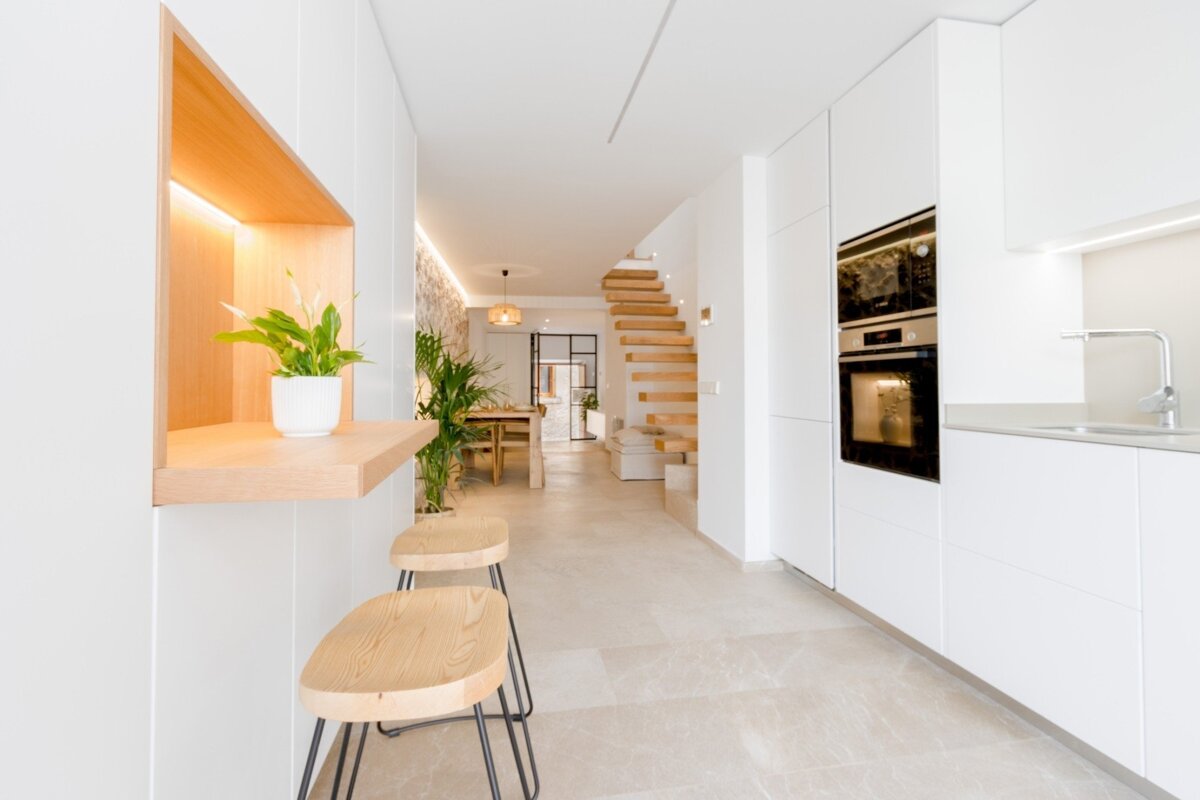 Modern, bright white kitchen with built-in oven, leading to a dining room and wooden stairs. Features a small breakfast bar with wood stools and plants.