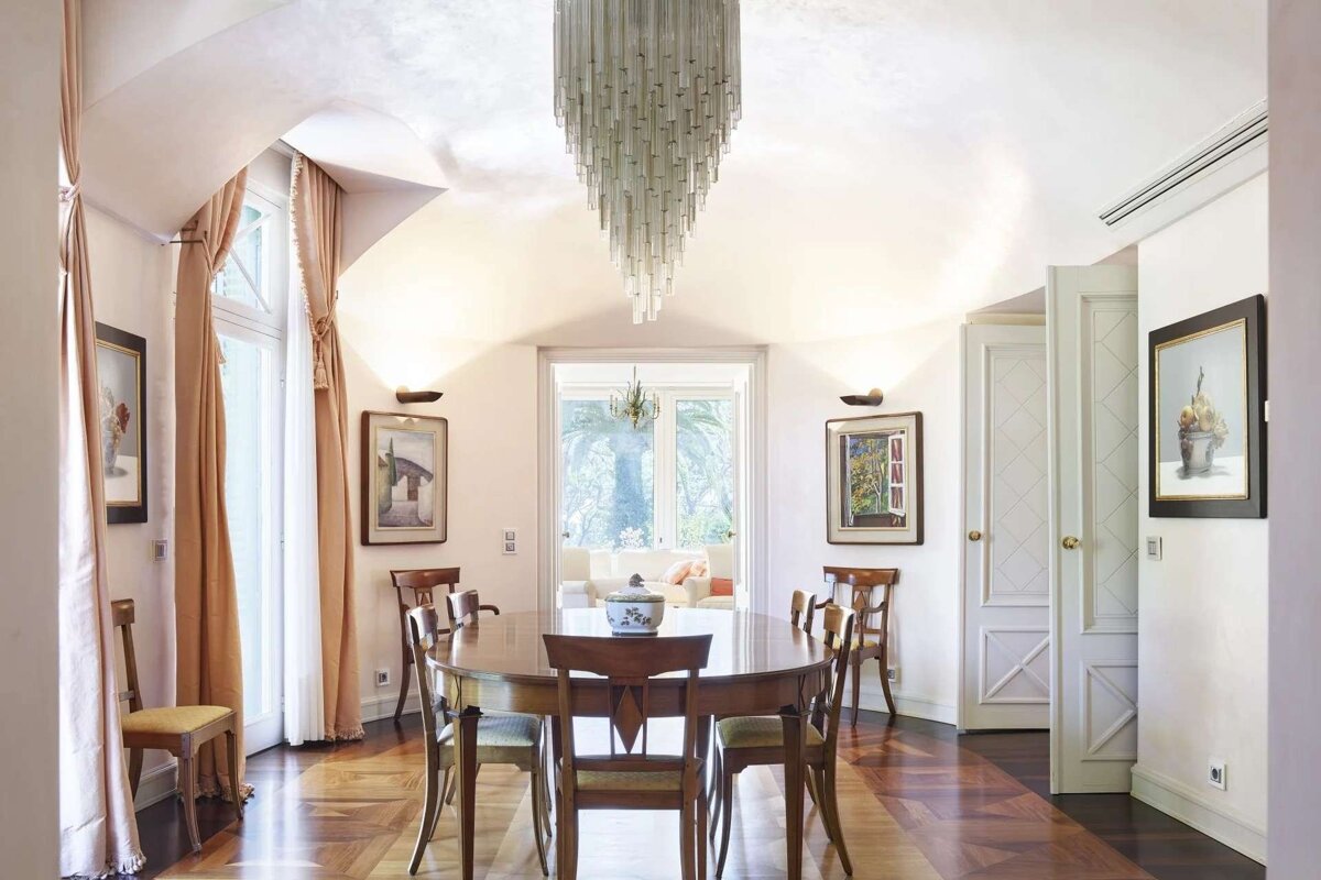 An elegant dining room features a large oval wooden table with chairs, a unique chandelier, and bright tall windows overlooking a garden.