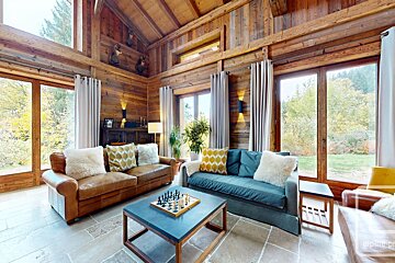 A cozy, rustic wooden living room with large windows framing nature views. Features comfy sofas, stone floors, and a chessboard on the coffee table.