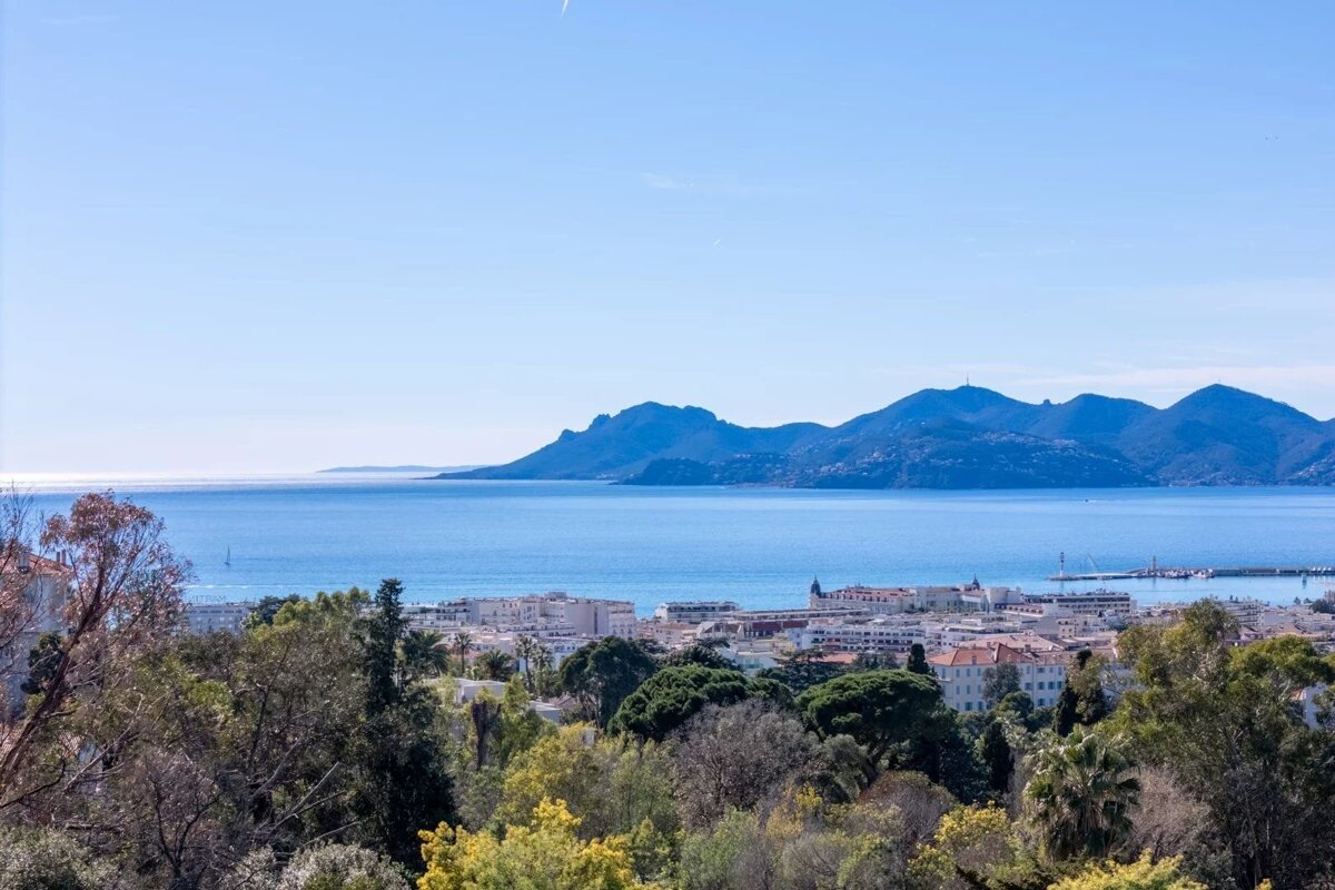 A scenic view of a coastal town along a blue sea, with green trees in the foreground and rolling mountains in the background.